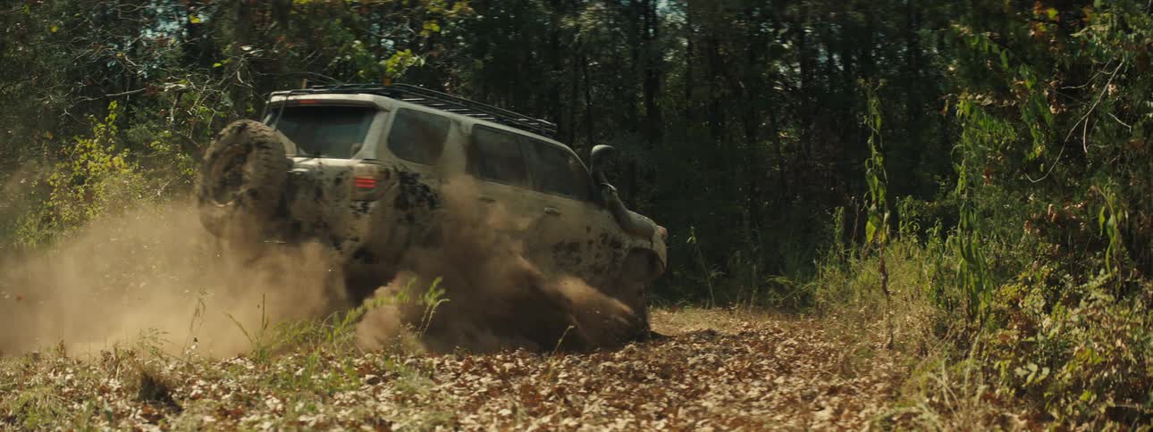 A tracking shot shows a tan vehicle covered in mud as it drives through a clearing in a forest while kicking up a large cloud of dust from the leaf covered ground