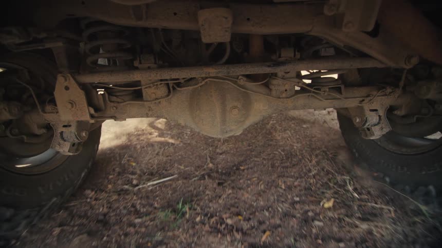 A lowangle view of a vehicles undercarriage as it moves slowly over dry uneven groundvisible components include the rear axle suspension springs and large offroad tires rotating on either side of the frame