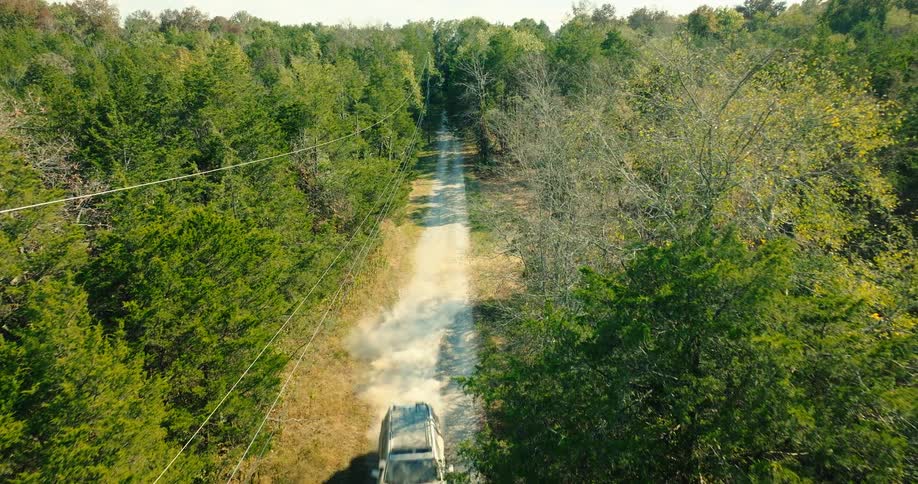 An suv driving out of a forest onto a dirt road in the countryside at midday with power lines and trees lining the road