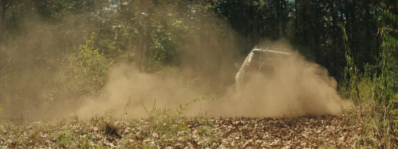 A tan muddy suv speeds along a narrow dirt path through a sunlit forest kicking up a thick cloud of dust as it disappears into the dense green trees and shadows