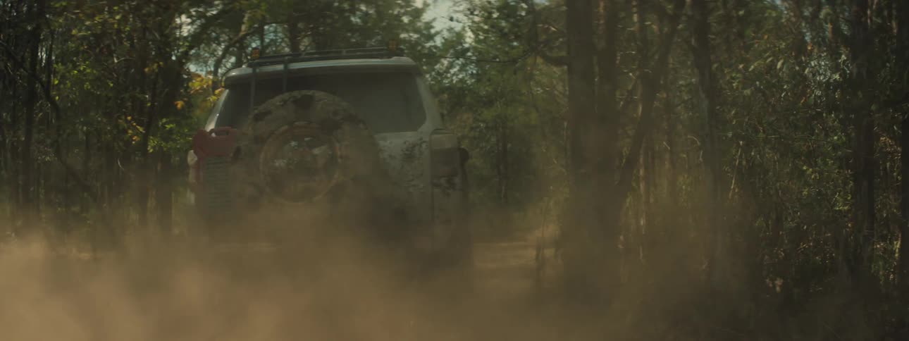 A tan mud covered vehicle with a roof rack and snorkel drives through a forest while throwing up thick clouds of dust