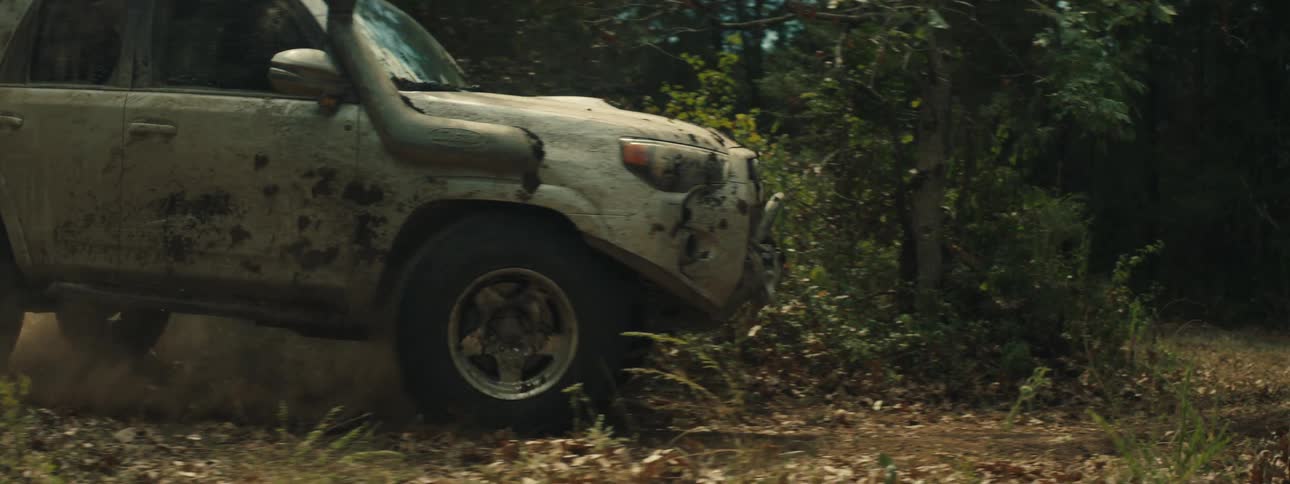 A muddy offroad suv speeds across a clearing in the woods kicking up a thick cloud of dust and dead leaves from the ground as it passes through the frame
