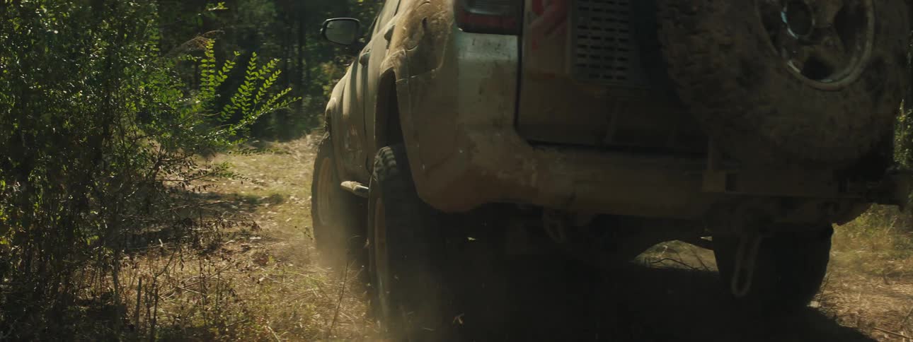 A car driving on a muddy road in the forest its body covered in mud and dust as it moves along the dirt road blowing dust behind it at midday