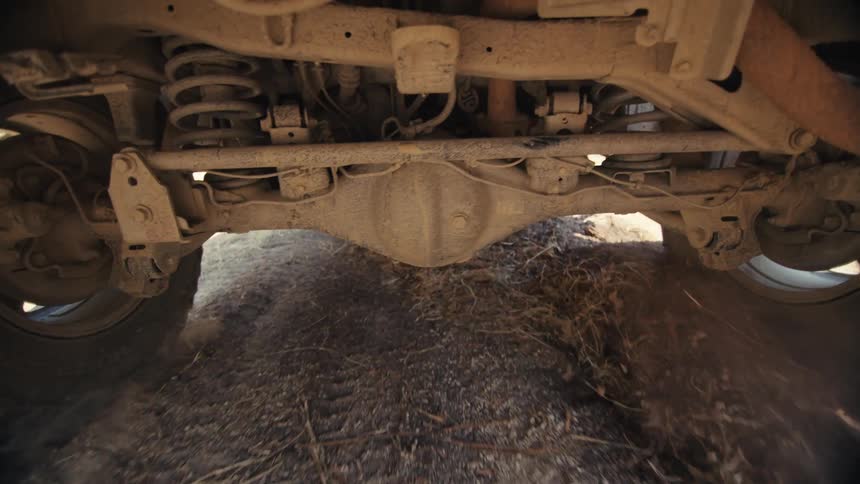 A low angle view from beneath a moving truck as it drives over a dry dirt trail showing the rear axle and suspension components bouncing while dust kicks up from the ground between the spinning tires