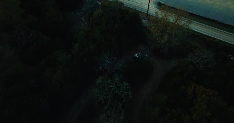 An overhead aerial shot shows a vehicle driving along a winding dirt path through a dense dark forest at twilight the cars headlights illuminate the surrounding trees as it navigates the shadows toward a nearby paved road