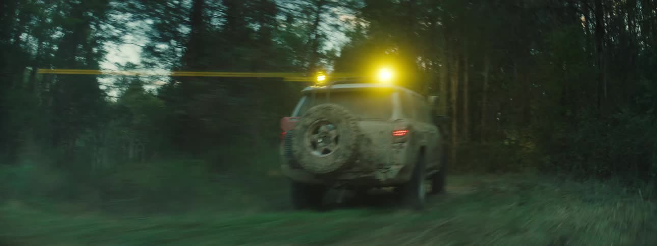A tan offroad suv speeds down a narrow dirt path through a dense forest at dusk kicking up thick clouds of dust while its yellow roof lights shine brightly and its rear tires bounce over the uneven terrain