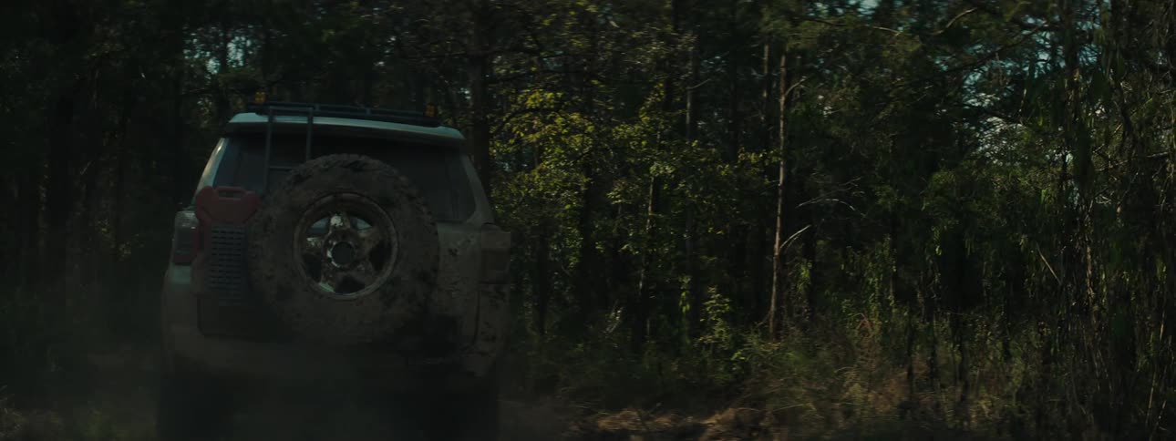 A view from behind shows a tan vehicle covered in thick mud driving away through a dark forest while throwing up dust