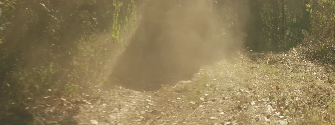 A tan off road vehicle drives away down a narrow dirt trail through a dense forest while kicking up a thick cloud of dust in the sunlight