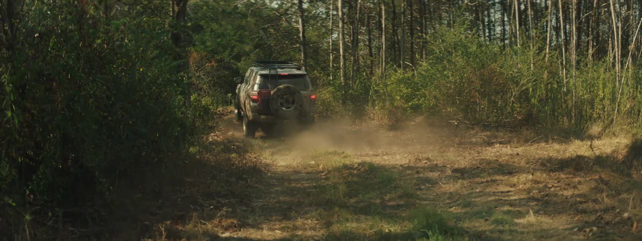 A view from behind shows a tan vehicle driving through a forest and over small bumps while leaving a trail of dust