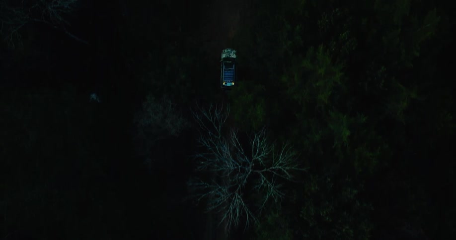 N aerial view shows a car driving along a narrow dirt road through a dark dense forest