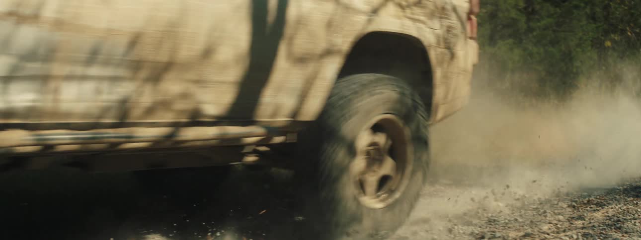 A tan offroad vehicle speeds down a narrow gravel road through a dense forest the truck kicks up a thick cloud of dust that lingers in the air as it passes the camera