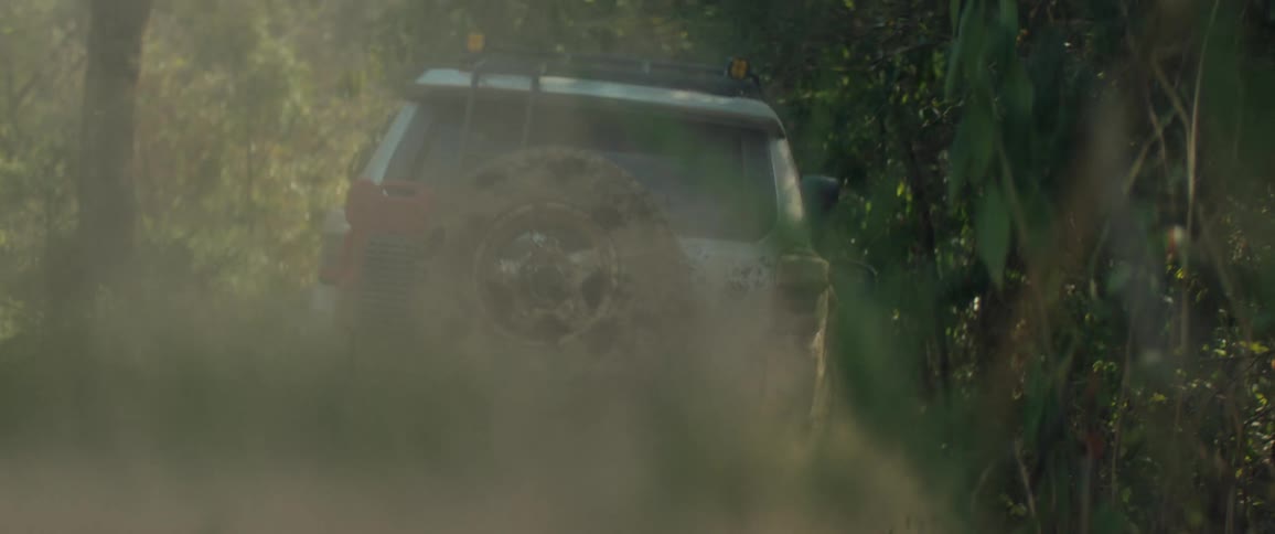 Mudcovered suv moves along a narrow trail between vegetation raising dust