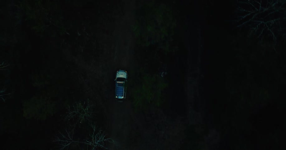 A top down aerial view shows a tan vehicle driving through a dark forest at night as it follows a winding dirt road