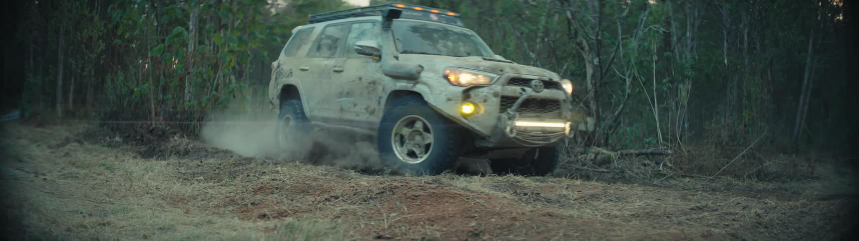 Toyota suv with headlights on drives along a dirt road in a forested area kicking up dust