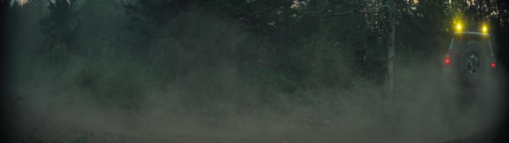 A tan offroad vehicle drives quickly down a dirt path through a forest at dusk