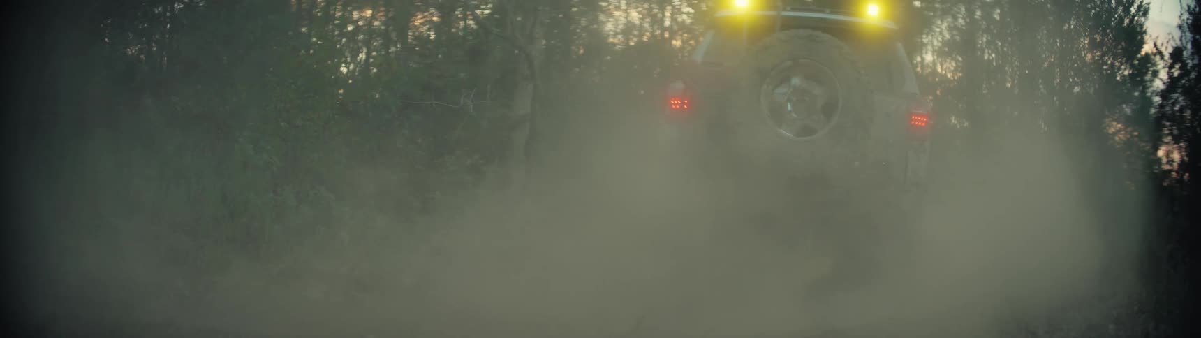 Suv with rear lights and roof lights on moves along a dirt road raising dust among trees