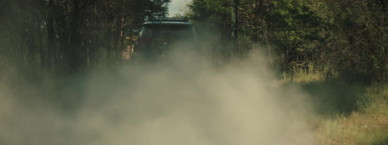 A shot from behind shows a mud covered vehicle as it turns onto a gravel road and speeds away while creating a massive cloud of dust that fills the air