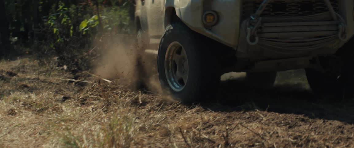 An offroad vehicle drives through a wooded area and kicks up a large cloud of dust the camera follows the front tires closely as they churn through the dry grassy terrain