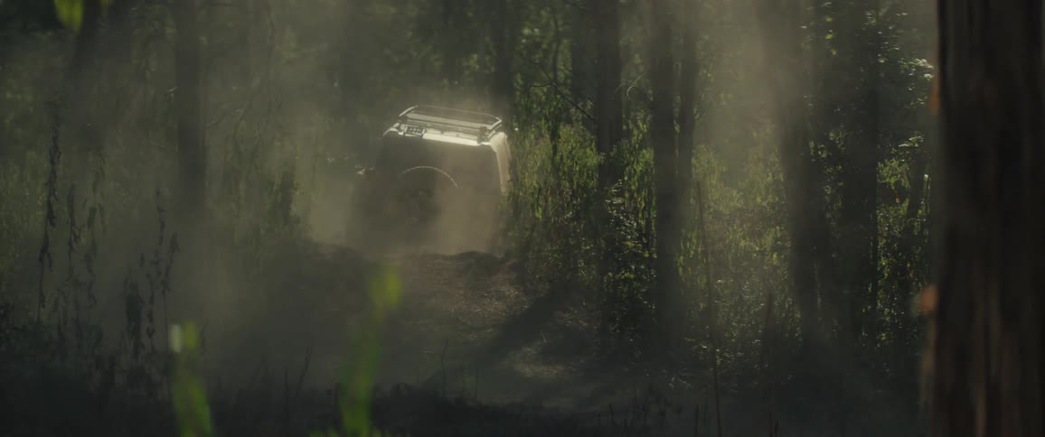 A view from behind shows a tan vehicle driving through a dense forest as sunlight filters through the trees and illuminates a thick cloud of dust