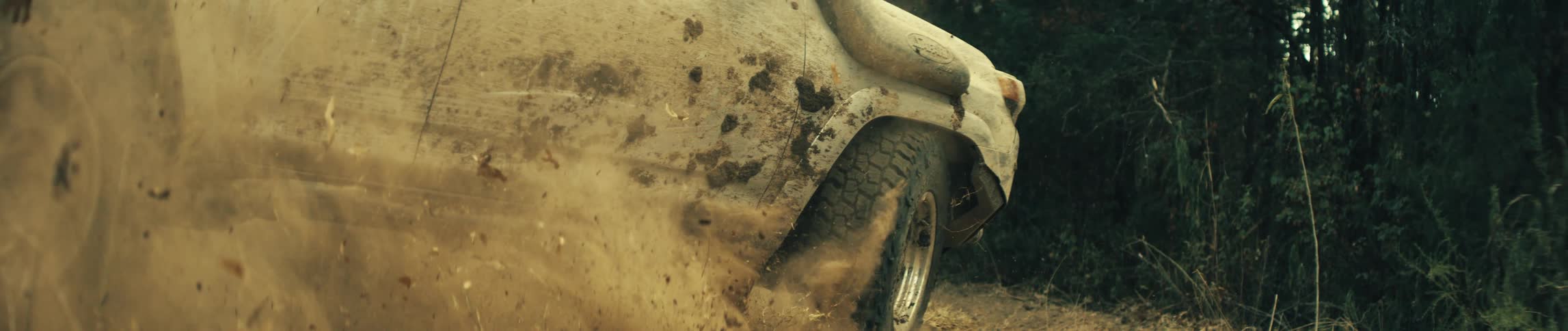 A muddy white offroad vehicle accelerating through a field its tires spin rapidly kicking up a large cloud of dust dirt and dried leaves