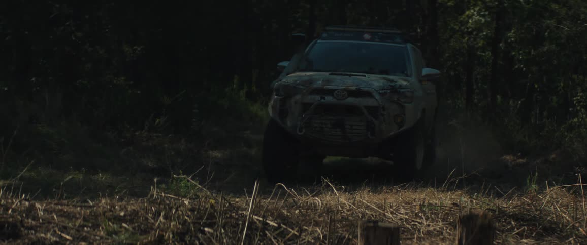 A silver offroad vehicle covered in mud drives out from a dark forest onto a sunlit clearing filled with dry grass and tall trees