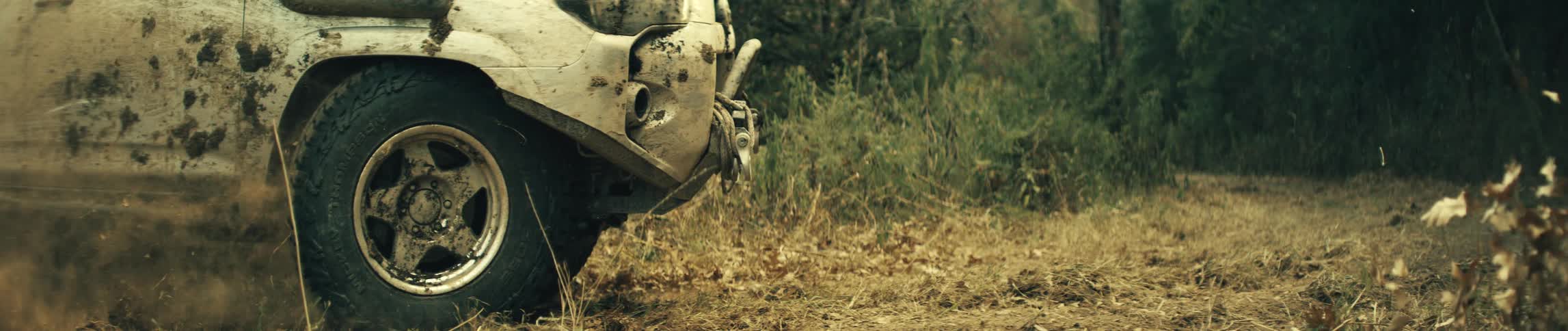 A muddy white suv car drives through a dry field of grass and fallen leaves