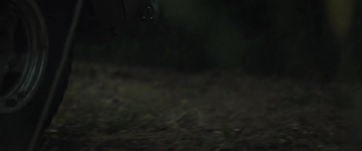 A tan vehicle with a roof rack and mud on the front drives through a dark forest and kicks up a thick cloud of dust