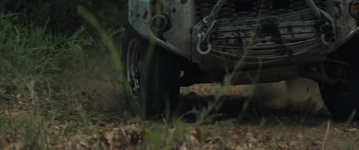 A tan mudsplattered toyota suv drives quickly down a narrow dirt path through a wooded areathe vehicle kicks up a thick cloud of dust and dry leaves as it passes closely by the camera