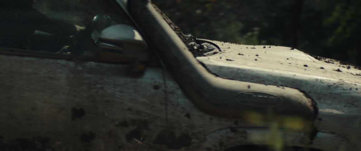 A tan mud covered vehicle with a roof rack and snorkel drives quickly past foliage while kicking up a thick cloud of dust