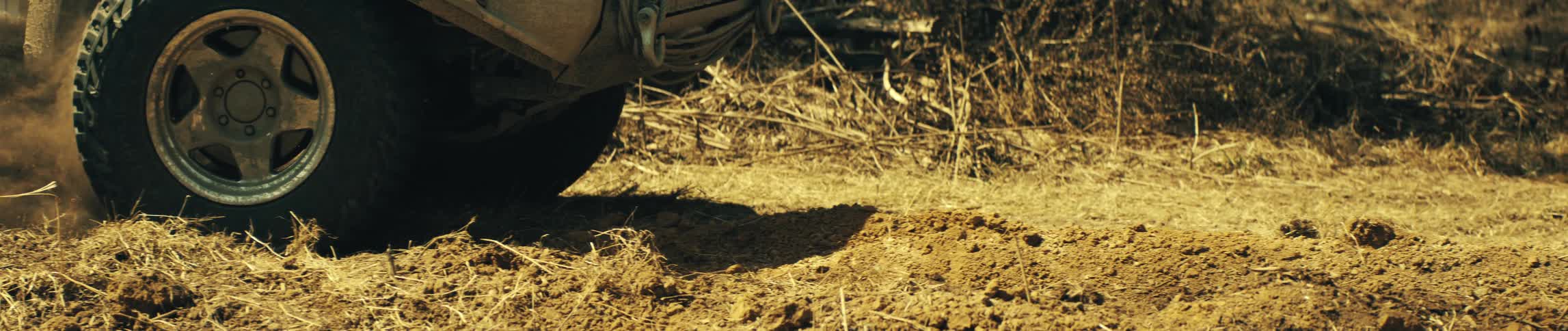 A close ground level view shows the front tire of a tan vehicle slowly moving across dry grass and dirt