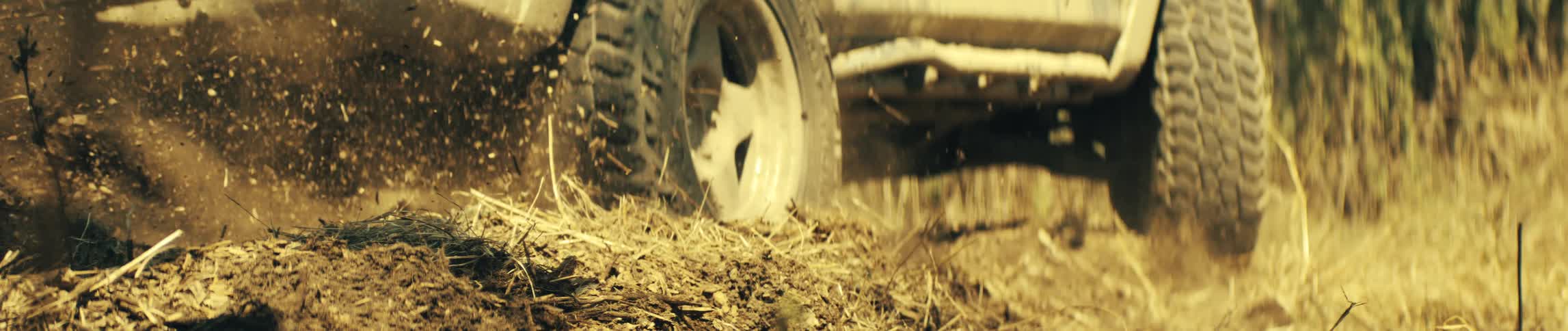 A close ground level view shows a large tire spinning and kicking up dirt and dry straw as the vehicle drives forward