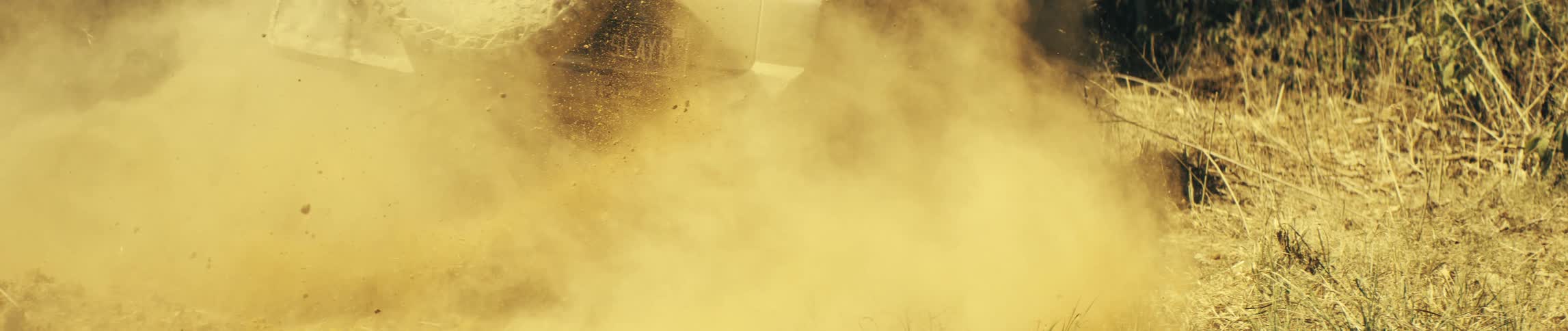 A vehicle speeds across a dry path kicking up a thick cloud of golden dust through the sunlit landscape