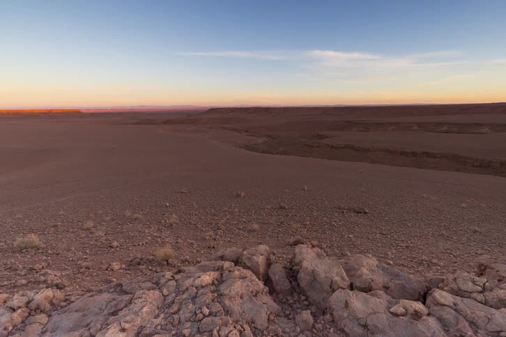 A vast rocky desert landscape as the sun rises casting a warm golden light across the arid terrain and distant plateaus the sky transitions from soft pink and purple hues to a clear blue highlighting the rugged textures of the foreground stones and th
