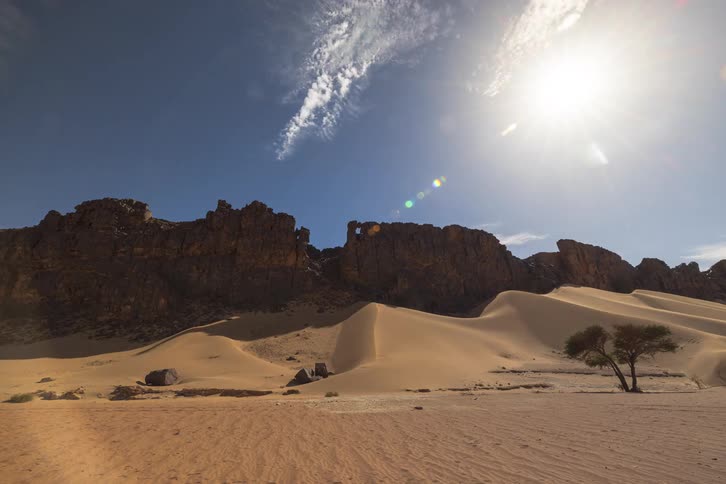 Clouds drift across the bright sun above a desert landscape featuring sand dunes and large rock formations