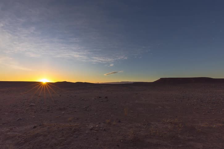 The sun rises over a vast rocky desert landscape casting warm golden light across distant hills beneath a wide clear blue sky