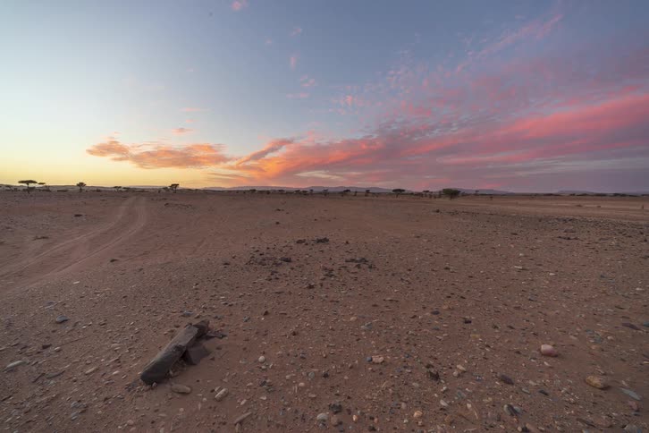 Dirt road crossing arid terrain with rocks and scattered trees under a sky with orange clouds at sunset