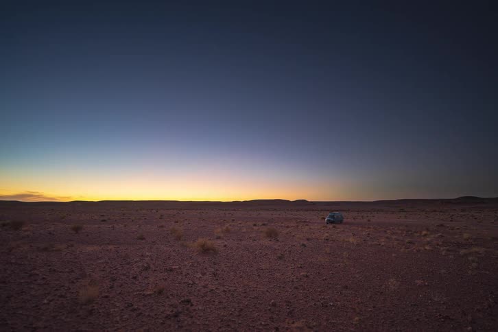 A van sits in a vast desert as the sun sets and stars appear in the deepening blue sky while headlights illuminate the surrounding arid ground and dry shrubs