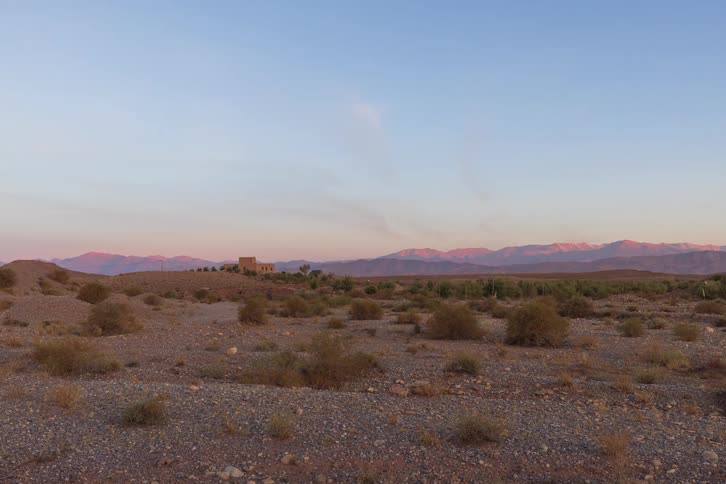 A dry rocky desert plain with sparse shrubs and a distant building against a mountain range