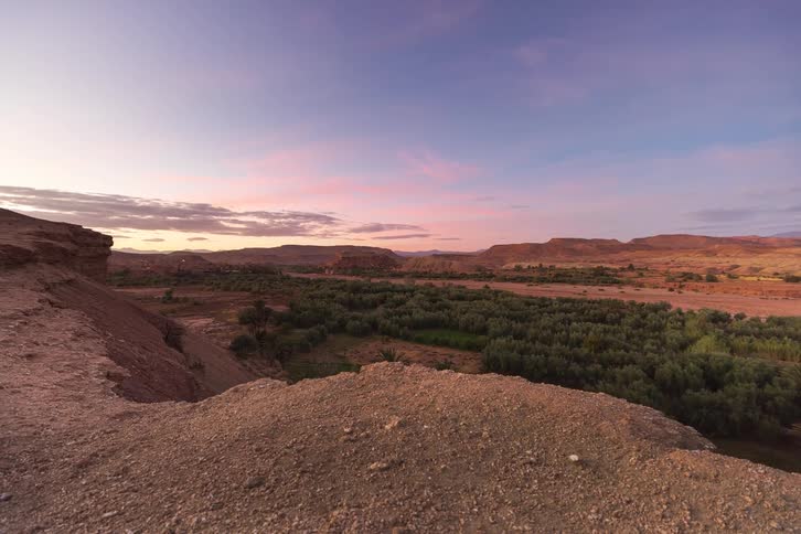 The camera pans over a rugged stone ledge overlooking a lush green oasis nestled within a vast desert valley under a vibrant sunset sky filled with wispy pink clouds and a fading purple light