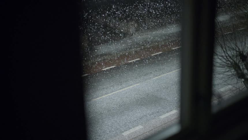 Snow falls steadily over a quiet dark road viewed through a window at night the streetlights reflect off the wet pavement as the winter flurries fill the air