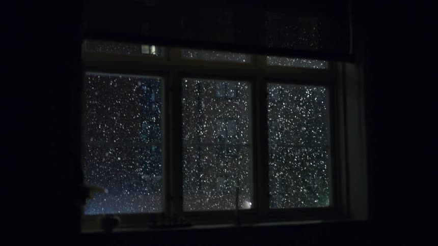 Snow falling outside a glass window viewed from indoors at night