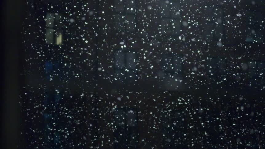 Snow falls slowly through the air between apartment buildings at night