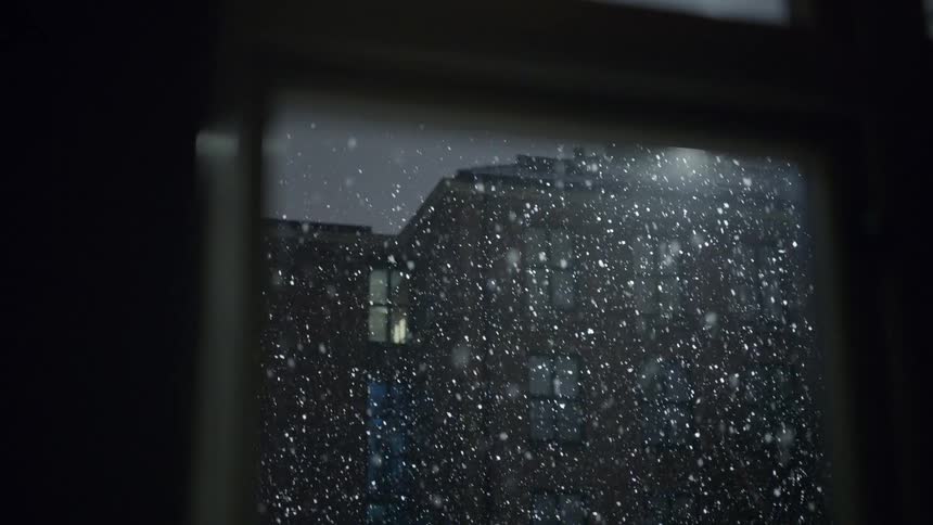 Snowflakes fall in front of a building seen through a window