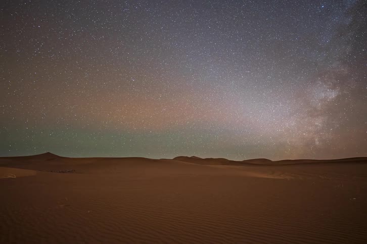 This timelapse video captures the milky way drifting across a vast starry sky above the silent undulating dunes of a desert as the night progresses the celestial band shifts and fades while a soft glow begins to emerge on the horizon