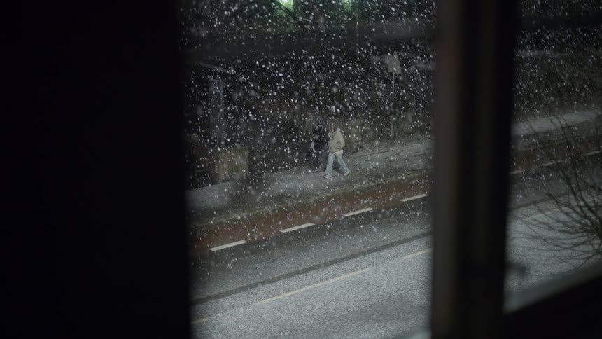 Pedestrians walking on a snowy sidewalk seen through a window frame at night