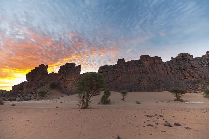 Towering rock formations rise from the quiet desert sands as the golden sky of sunset glows behind them