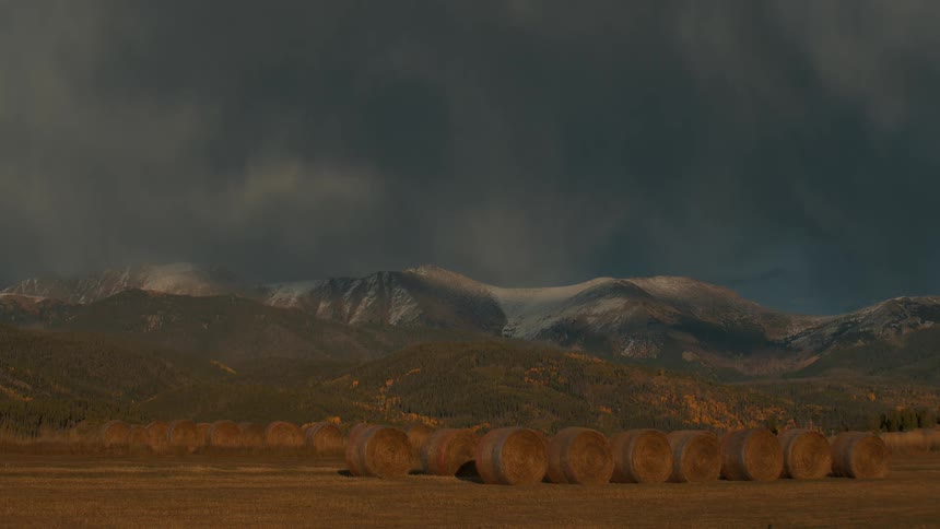 A wide shot of a rural landscape under a heavy dark storm cloud in the foreground several large hay bales sit in a harvested field while a mountain range with dusted snow peaks stands in the distance