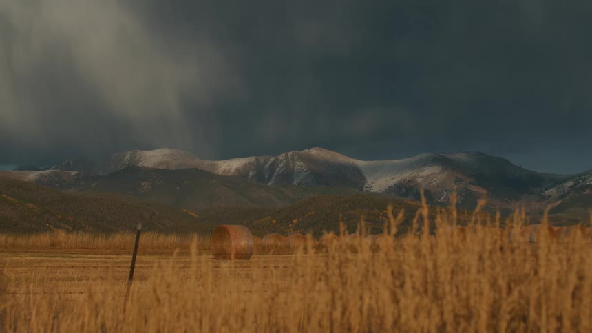 A dramatic wideangle landscape where the horizon is split by two distinct layersin the foreground a field of golden sunlit tall grass and large hay bales stretches across the bottom third of the frame above it a massive range of dark rugged moun