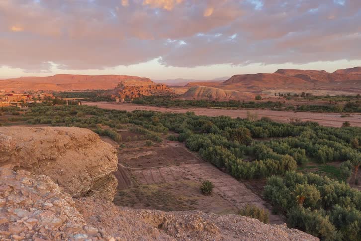 A wide shot shows the ancient earthen city of ait benhaddou nestled in a river valley as the rising sun casts a golden glow over green palm groves and barren mountains under a sky with purple clouds