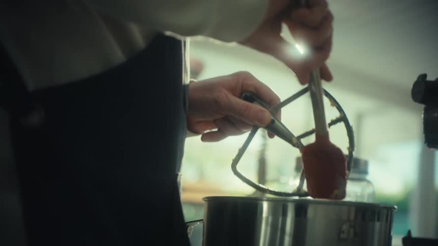 A baker scrapes thick dough from a stand mixer beater with a metal spatula over a stainless steel bowl in a kitchen